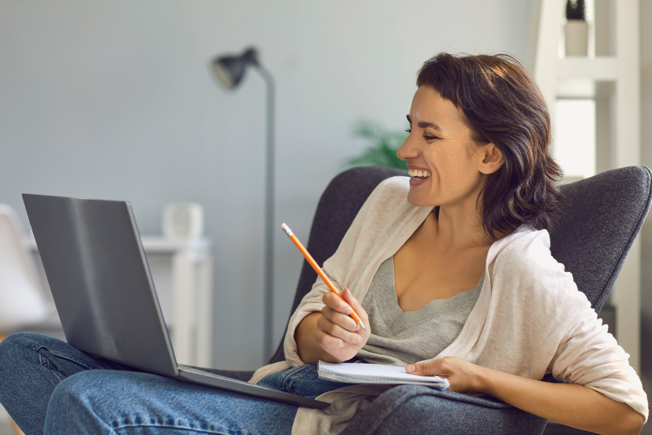 Smiling Woman Sitting with Laptop and Making Notes during Online Meeting